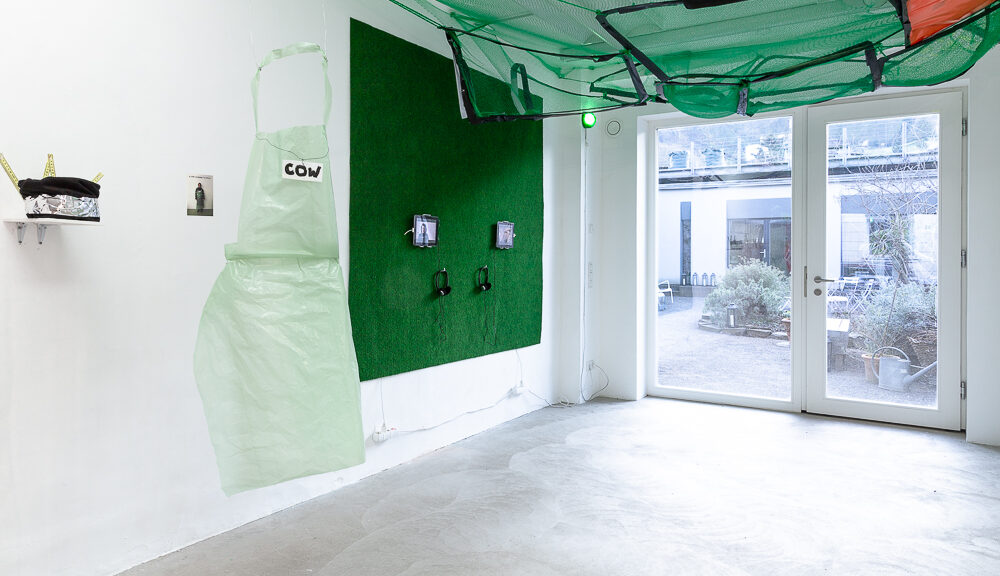 Ausstellungsansicht, grünes Netz an der Decke, grüner Teppich mit zwei Bildschirmen und Kophörer an der Wand befestigt, Kunststoffolien Schürze