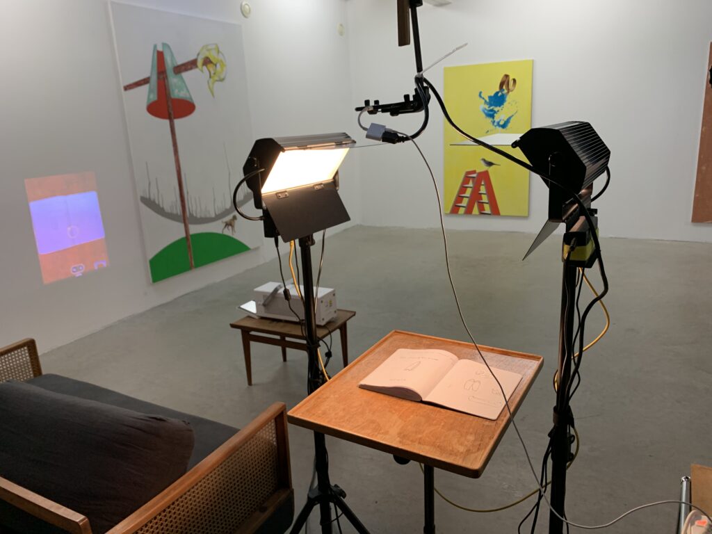 Exhibition view with two large acrylic on canvas and projection of the installation, table sketchbooks and lamps