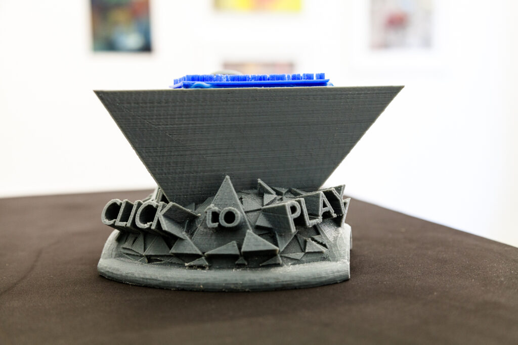 3D printed Objekt grau mit Aufschrift "click to play", auf schwarzer Oberfläche