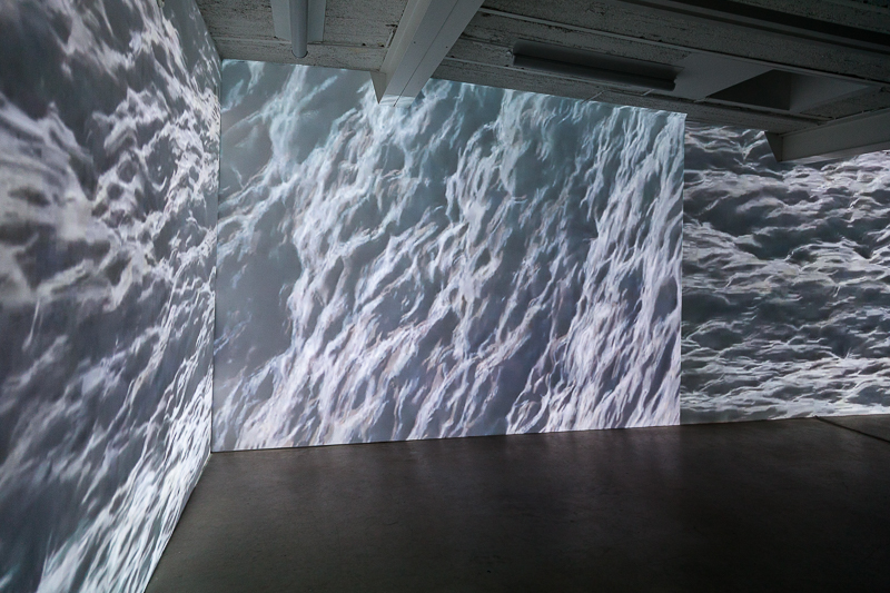 Eine Aufnahme des Ausstellungsraumes, auf die weißen Wände werden Nahaufnahmen von Wellen projeziert. Die Projektion nimmt die gesamte Wandfläche ein.