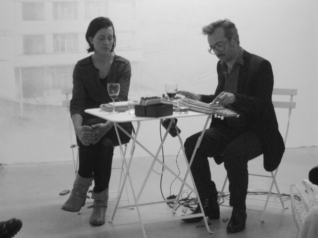 Zwei Personen sitzen an einem Tisch, die rechte Person scheint an einem Instrument etwas einzustellen, die linke Person schaut ihr dabei zu.